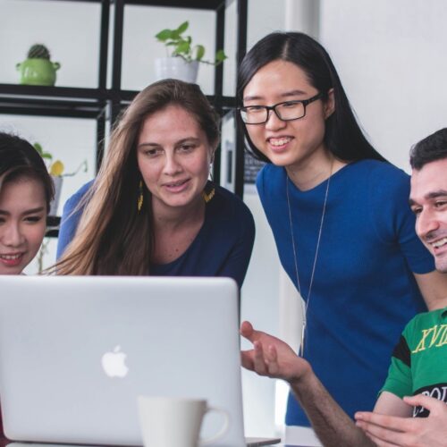 Multiracial team leaning in around a laptop, exchanging ideas with engaged expressions—representing inclusive collaboration, strategic alignment, and the momentum of group problem-solving.