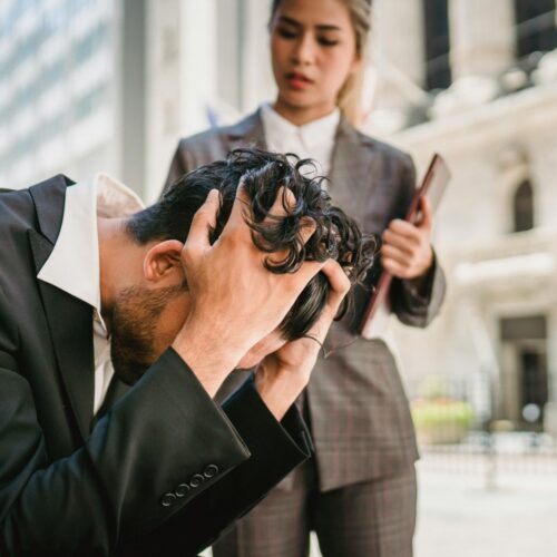Man sits with head in hands, visibly overwhelmed, as a colleague stands beside him—capturing the emotional weight of professional pressure and the potential for reflection, support, and reset.