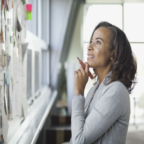 Woman standing in front of a wall covered in colorful sticky notes, holding a marker pen and gazing thoughtfully—capturing a moment of strategic reflection, emotional clarity, and creative planning